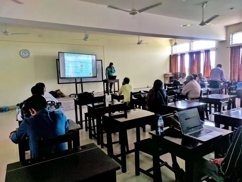 A presenter speaking in a classroom while participants listen and follow on laptops