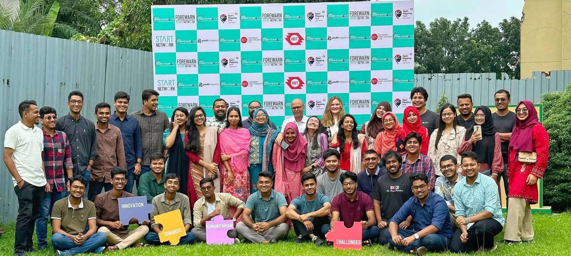 A large group of hackathon participants posing on the grass in front of a sponsor wall