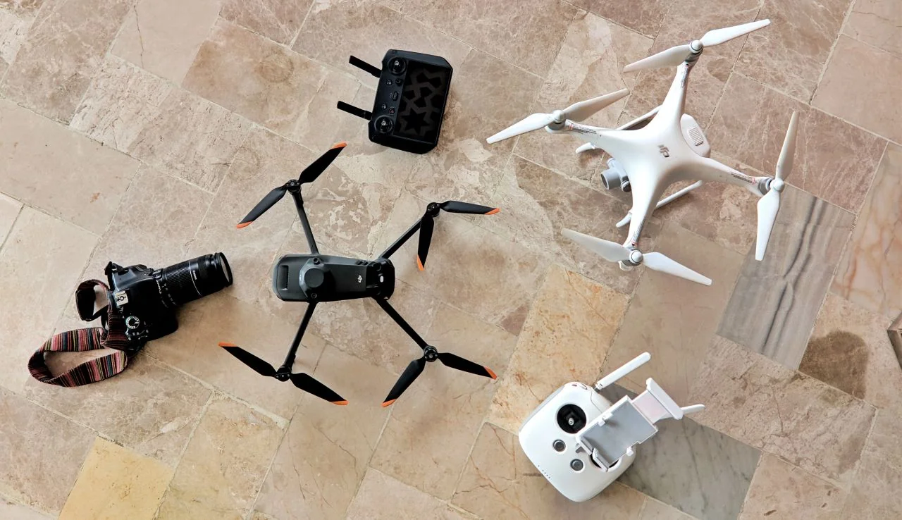 Two DJI drones, a black folding drone and a white quadcopter, with two remote controllers and a DSLR camera on a stone floor