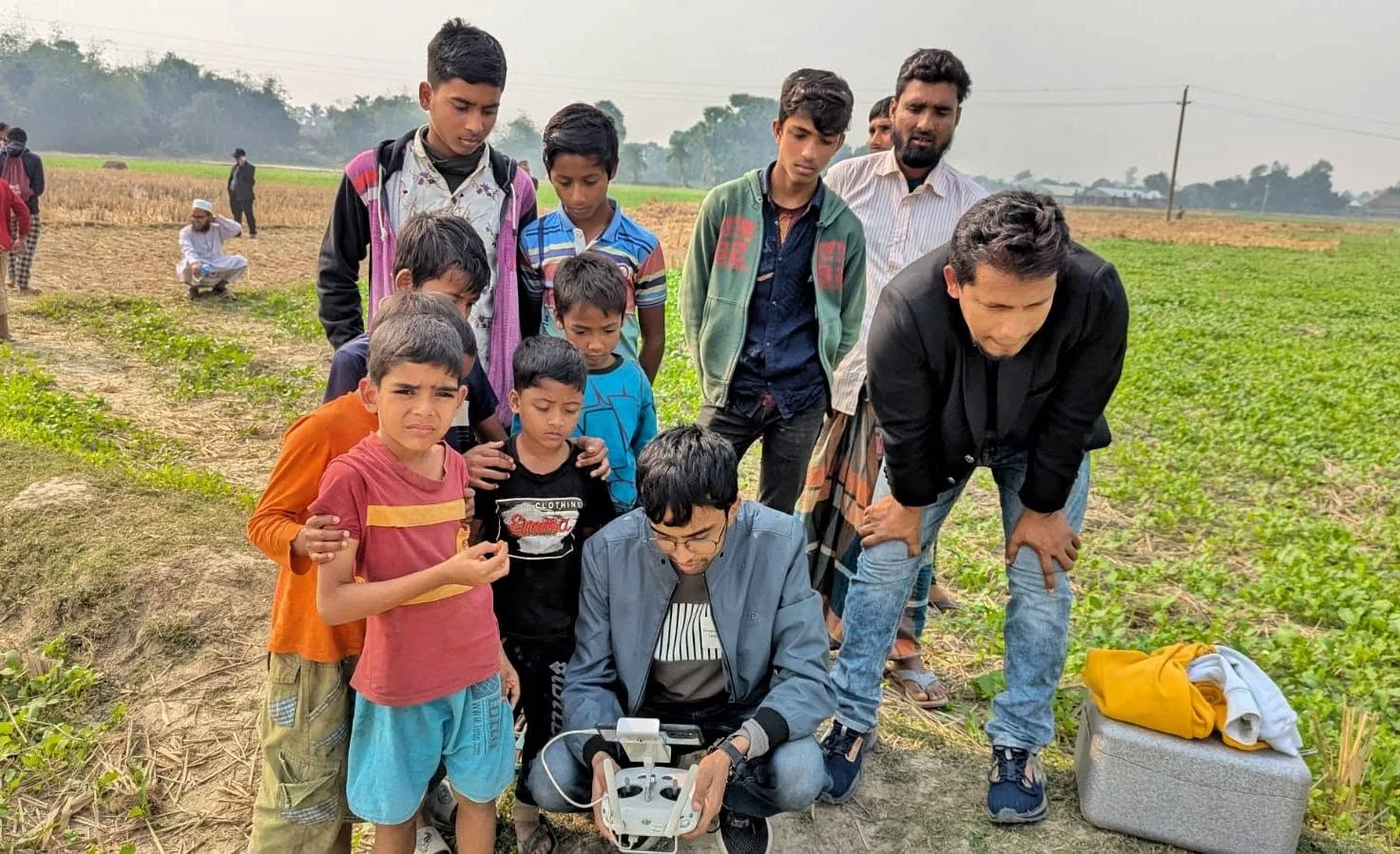 A young man crouching in a field with a DJI drone controller, surrounded by curious village children and adults